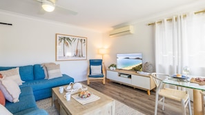A living room with a blue sofa, a wooden coffee table with a tray of food, a TV on a stand, and a chair with a cushion. - Sunset Shores 1 Surf Air , 24 Marine Dr - air con , Wi-Fi, across to the beach (Fingal Bay)