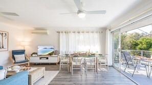 A modern living room with a flat-screen TV, a dining area with a glass table, and a balcony with outdoor seating. - Sunset Shores 1 Surf Air , 24 Marine Dr - air con , Wi-Fi, across to the beach (Fingal Bay)