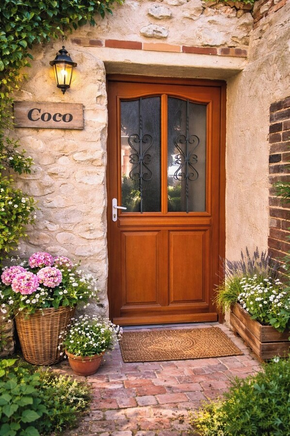 Una porta in legno con un vetro e un cancello in metallo, un muro in pietra con un cartello che riporta 'Coco' e piante in vaso con fiori.