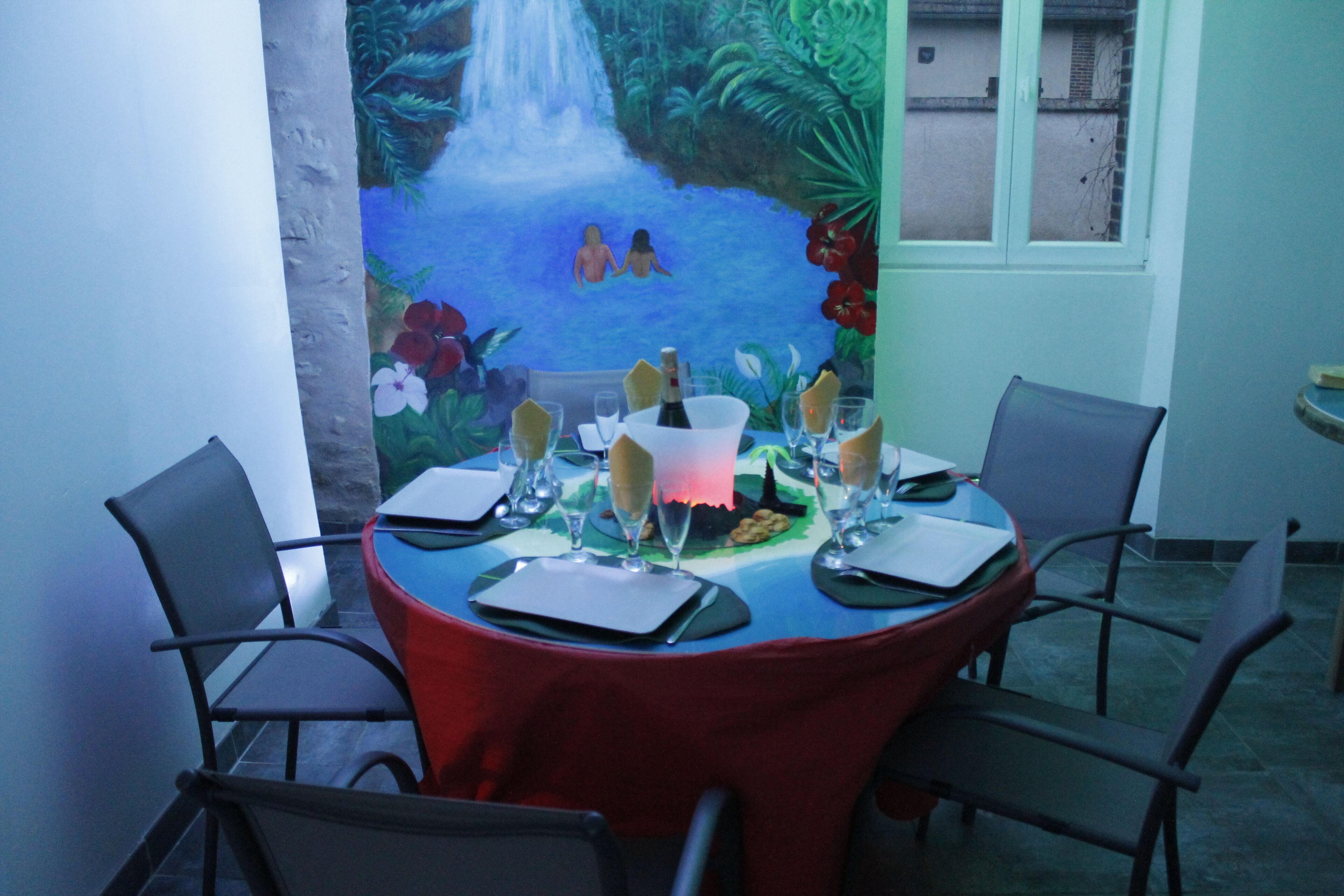 Een eettafel gedekt voor vier personen, met stoelen, een rode tafelkleed en een muurschildering van een waterval en tropisch landschap op de achtergrond.