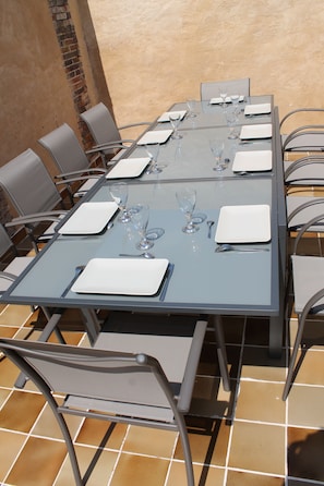 Un tavolo da pranzo rettangolare all'aperto con sedie in metallo, apparecchiato con piatti bianchi, bicchieri trasparenti e posate.
