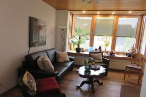 A living room with a black sofa, a coffee table, and a wooden chair. There is a window with blinds and a potted plant on the windowsill. - Holiday apartment in Konz with swimming pool (Konz)