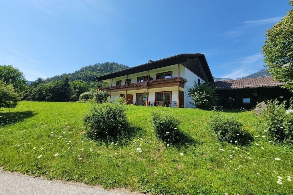 Un bâtiment à deux étages, doté d’un toit en pente, entouré d’un champ verdoyant parsemé de fleurs sauvages, avec une montagne en arrière-plan.