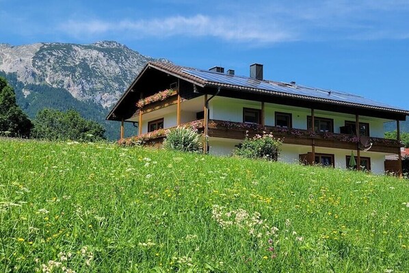 Un chalet de dos pisos con techo inclinado, paneles solares y un balcón con macetas de flores.