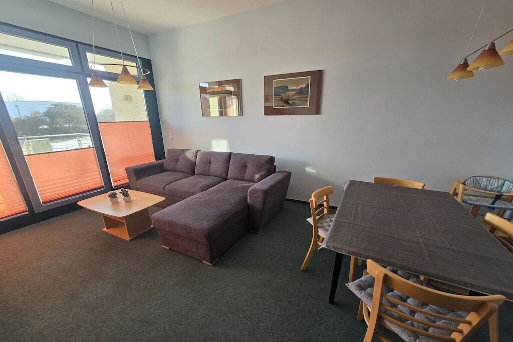 Una sala de estar con un sofá modular, una mesa de comedor con sillas y ventanas grandes con persianas.