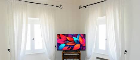 A room with two windows, white curtains, a television on a wooden stand, and a small white bench.