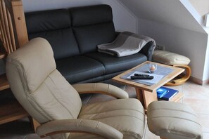 Un salon avec un canapé noir, des fauteuils beiges, une table basse en bois et une télécommande.