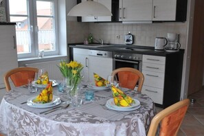Une table à manger dressée avec des assiettes, des couverts et des serviettes, un vase rempli de fleurs, et un coin cuisine avec des placards et des appareils électroménagers.