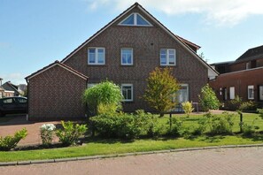 Une maison à deux étages, construite en briques, avec un toit en pente, une cour avant clôturée et une voiture garée dans l’allée.