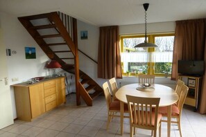 Un comedor con una mesa redonda, sillas, una escalera de madera, un televisor y una ventana con cortinas.