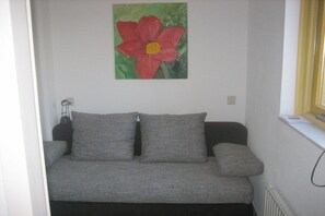 A grey sofa with two cushions against a white wall, a painting of a red flower above it, and a window with a yellow blind to the right. - Holiday home with 3 bedrooms (PD Julianadorp)