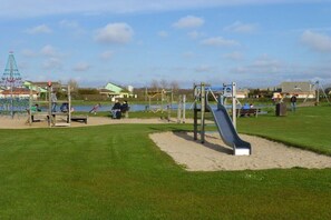 A playground with a slide and swings, a small boat, and houses in the background. - Holiday home with 3 bedrooms (PD Julianadorp)