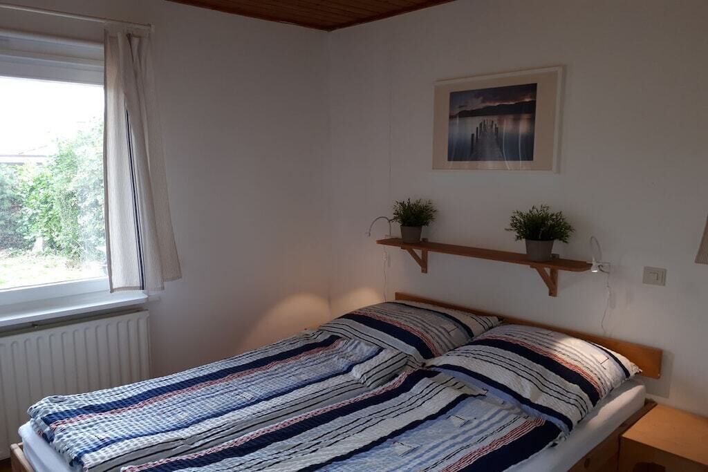 A bedroom with a bed, a window with curtains, a radiator, a wall-mounted shelf with two potted plants, and a framed picture.