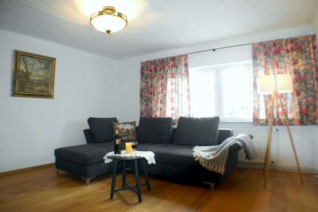 Een kamer met een donkergrijze bank, een klein tafeltje met een fles en een kaars, een schilderij aan de muur en een raam met bloemen gordijnen.