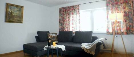 Een kamer met een donkergrijze bank, een klein tafeltje met een fles en een kaars, een schilderij aan de muur en een raam met bloemen gordijnen.