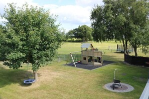 Ein Spielplatz mit Schaukeln, einem kleinen Holzspielhäuschen und einem umzäunten Bereich.