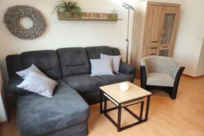 Una sala de estar con un sofá seccional gris, una mesita de centro de madera, un sillón gris, un mueble de madera y una planta colgada en la pared.