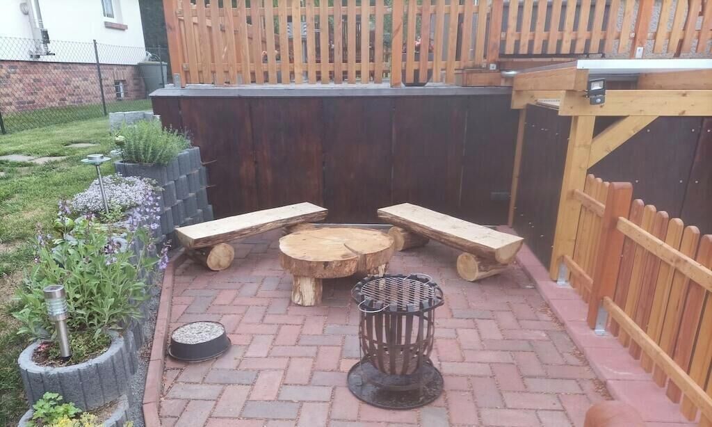 An outdoor patio with a wooden table and benches, surrounded by a brick floor and a wooden fence.