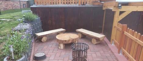 An outdoor patio with a wooden table and benches, surrounded by a brick floor and a wooden fence.
