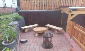 Een terras buiten met een houten tafel en bankjes, omringd door een betegelde vloer en een houten hek.