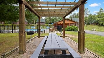 A wooden pergola with picnic tables, a swimming pool, and a building in the background.