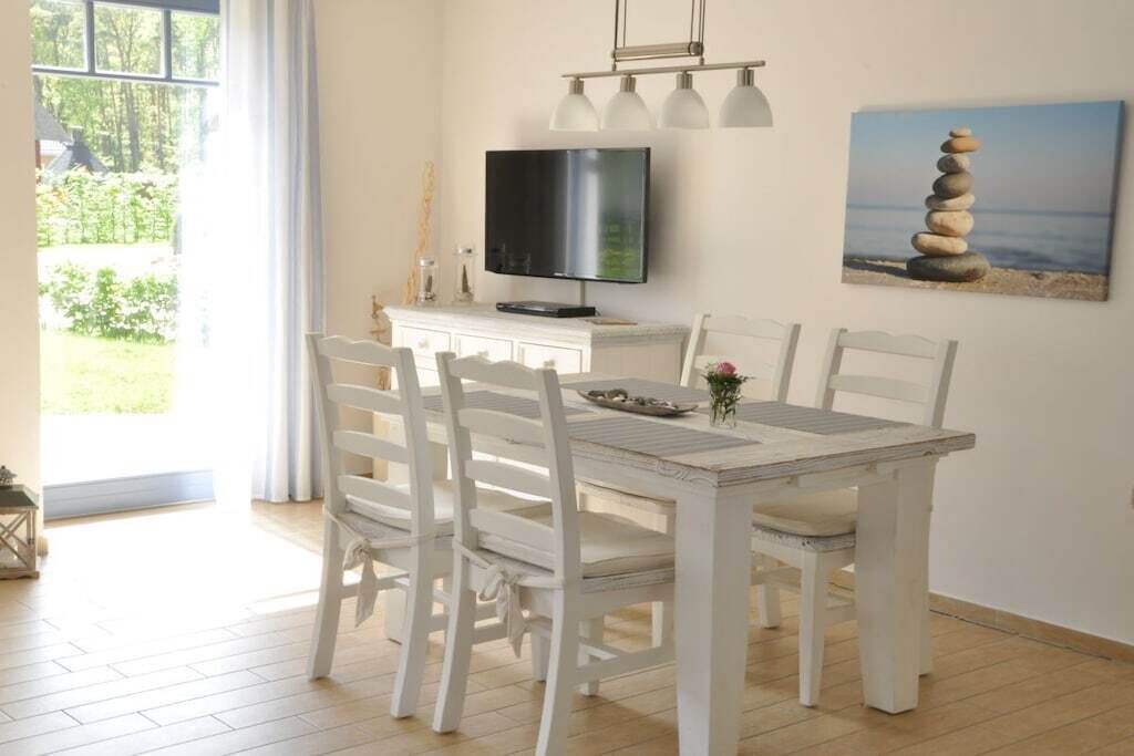 A dining area with a white table and chairs, a television on the wall, and a painting of stacked stones.