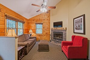 A cozy cabin interior with a wooden interior, a fireplace, a red armchair, and a ceiling fan.