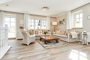 A spacious living room with a large sofa, a coffee table, and a wicker chair. There are windows with curtains, a painting on the wall, and a lamp on a side table. - Cozy "Wadden Sea gem" with south-facing terrace (Munkmarsch)