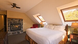 Une chambre avec une cheminée en pierre, un lit recouvert de draps blancs, un fauteuil rouge et un ventilateur de plafond.