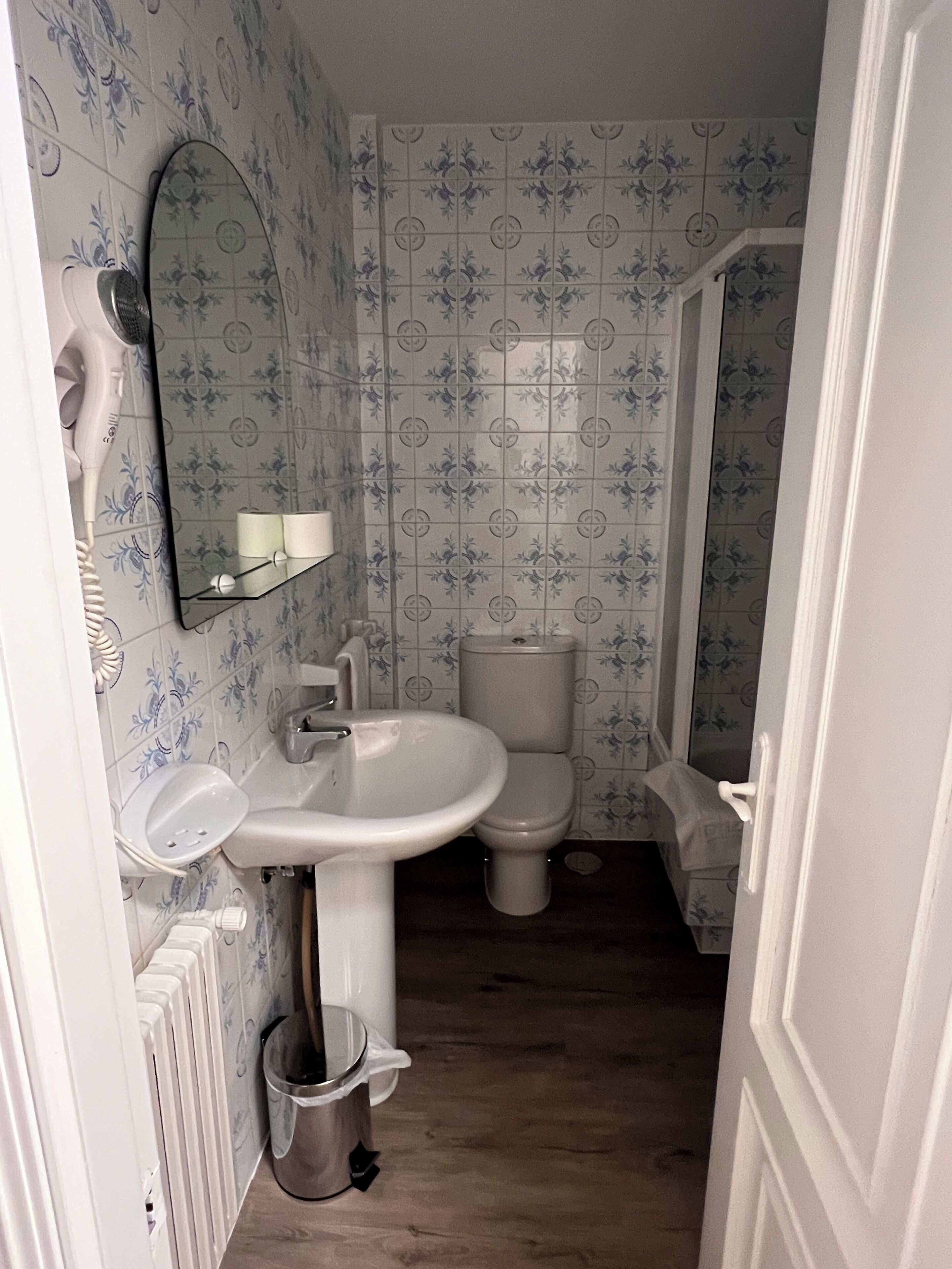 A bathroom with a white sink, toilet, and a mirror above the sink. There is a radiator on the left and a trash bin under the sink.