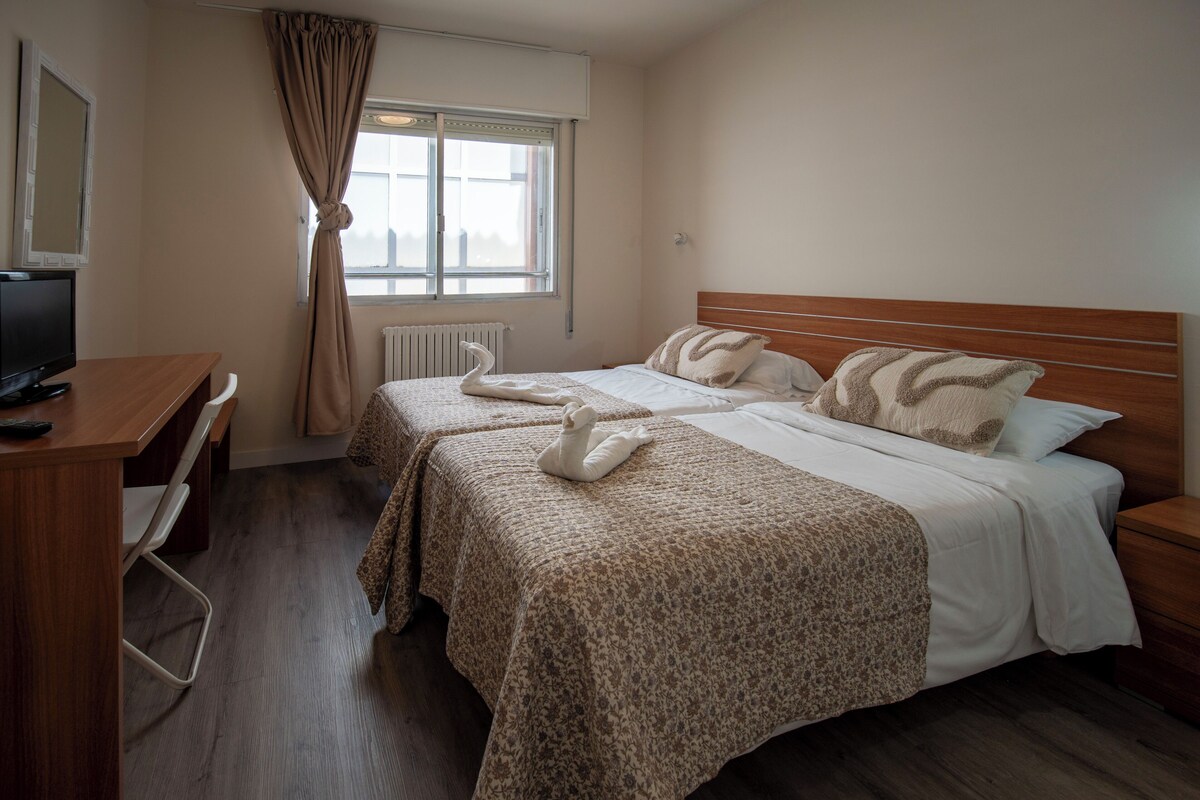 A hotel room with two beds, a desk, a chair, a television, and a window with curtains.