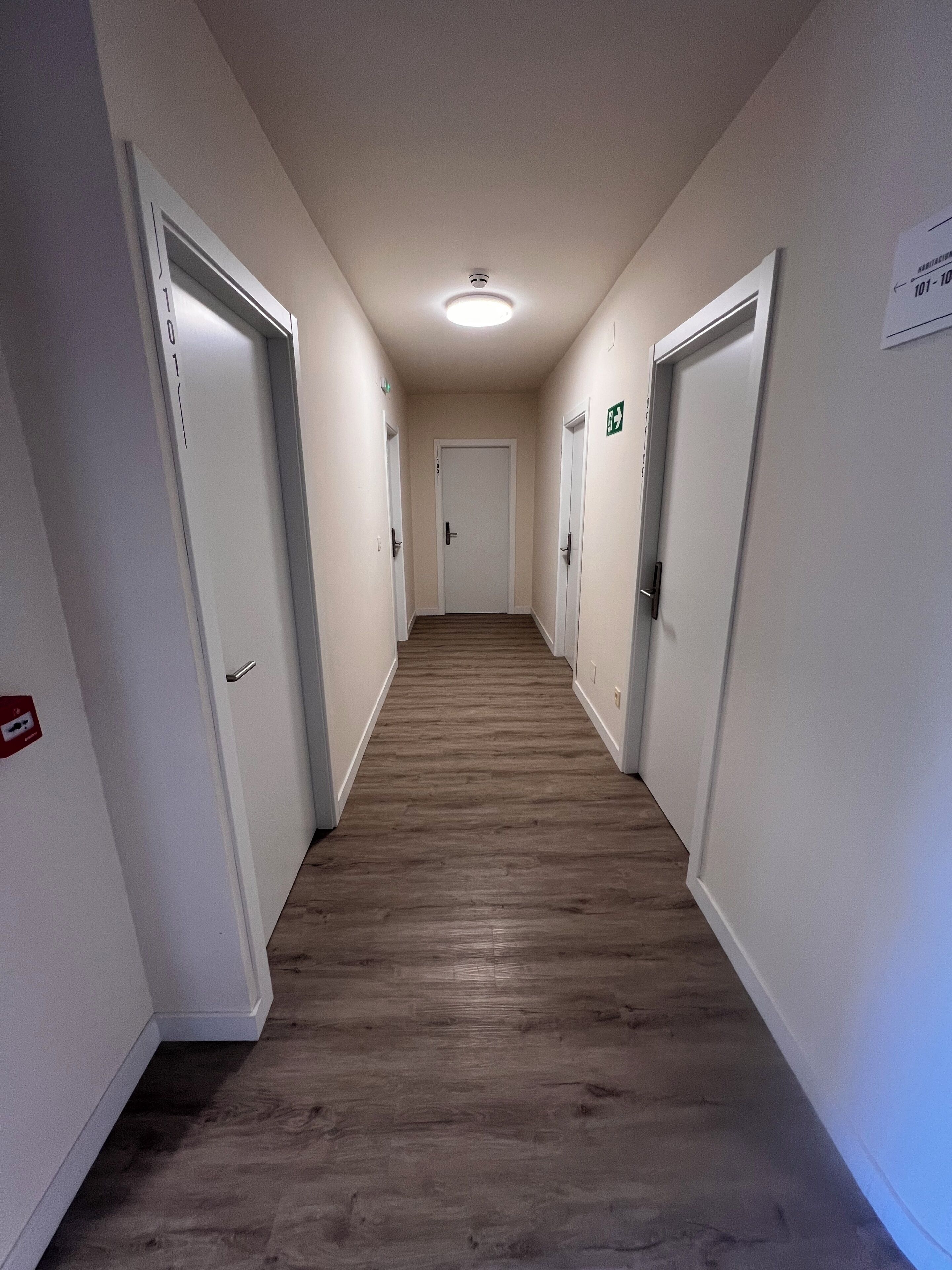 Un pasillo con puertas a ambos lados, suelo de madera y una lámpara en el techo.