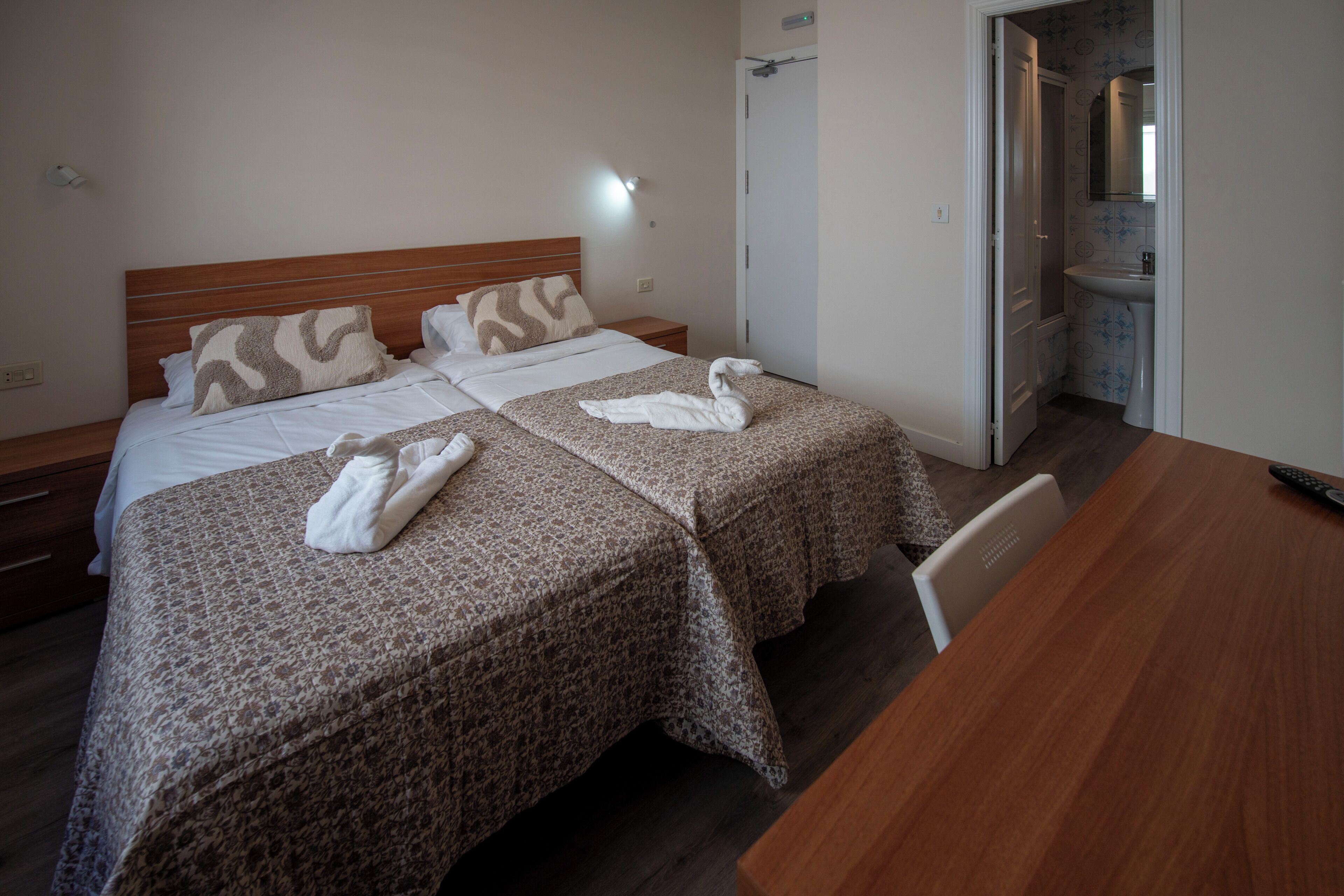 Habitació d'hotel amb dos llits, un panell de capçalera de fusta, una paret blanca, un bany visible a través d'una porta oberta i una taula de fusta.