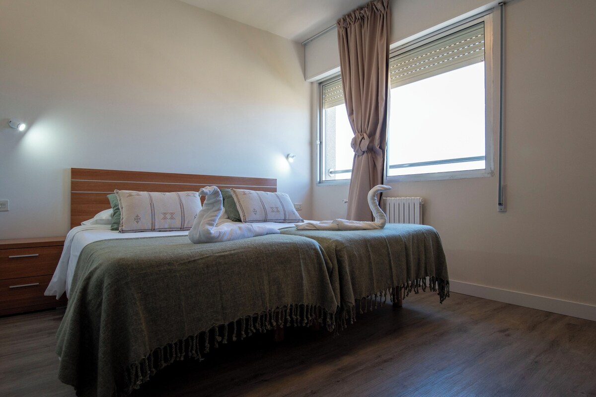 Een slaapkamer met een houten hoofdeinde, een bed met een groene deken, kussens en een kussen in de vorm van een zwaan. Er is een raam met gordijnen en een radiator aan de muur.