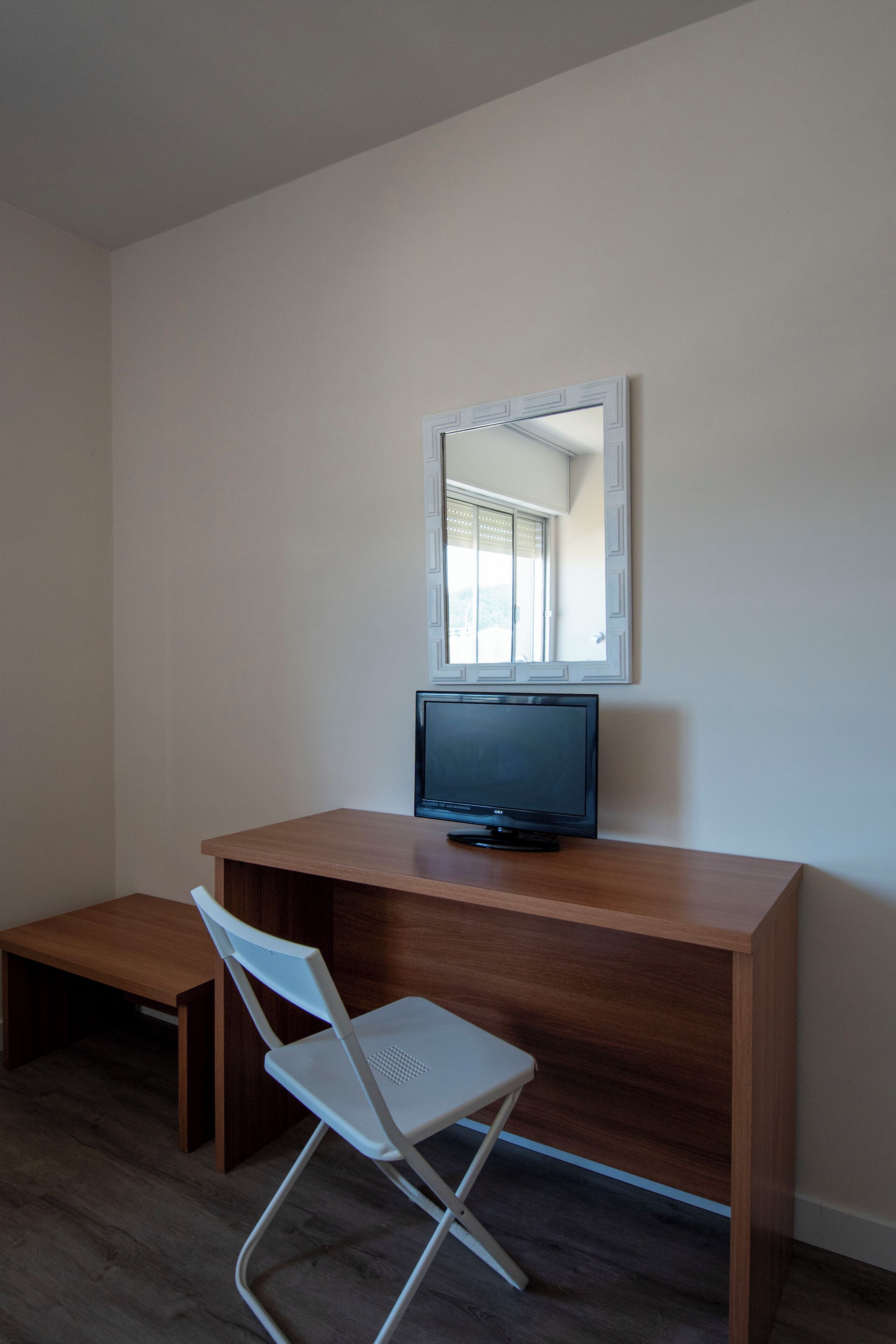 Habitació petita i moderna amb escriptori de fusta, televisió i cadira plegable blanca.