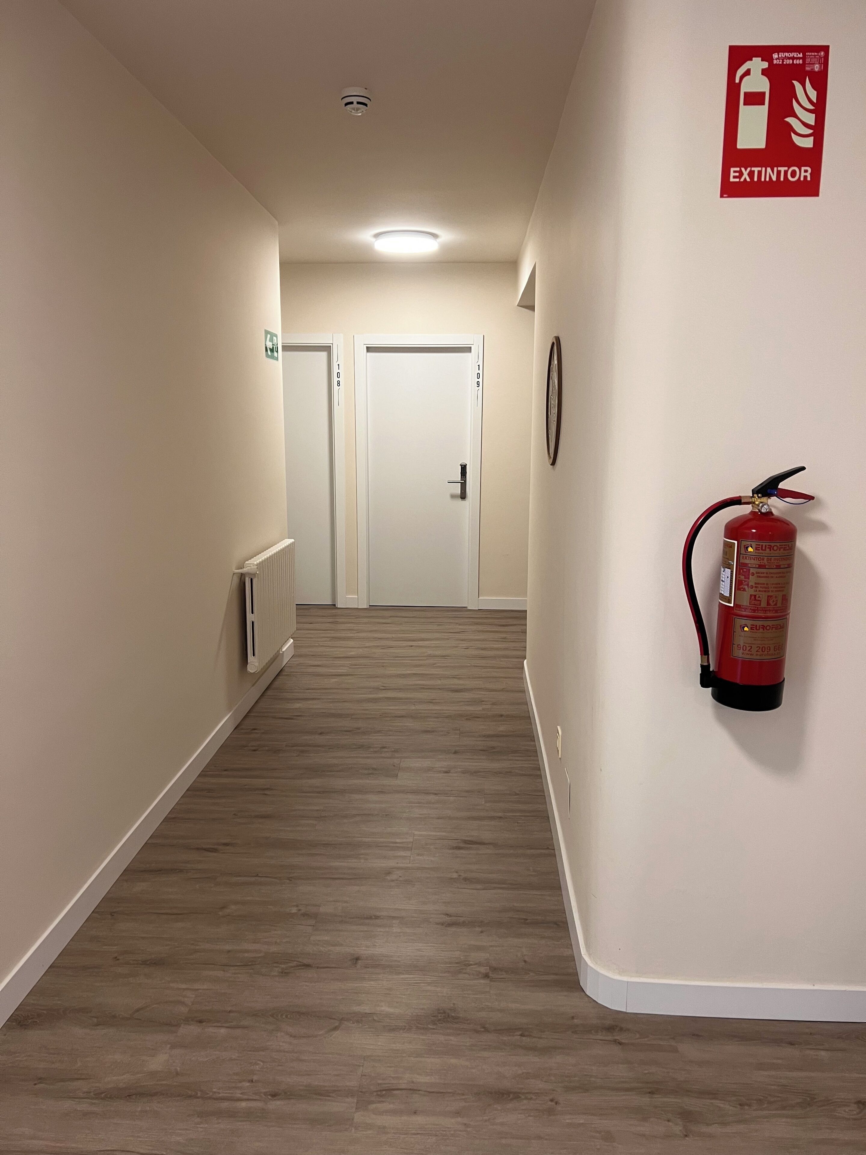 Un pasillo con un extintor montado en la pared, una lámpara en el techo y puertas al final.