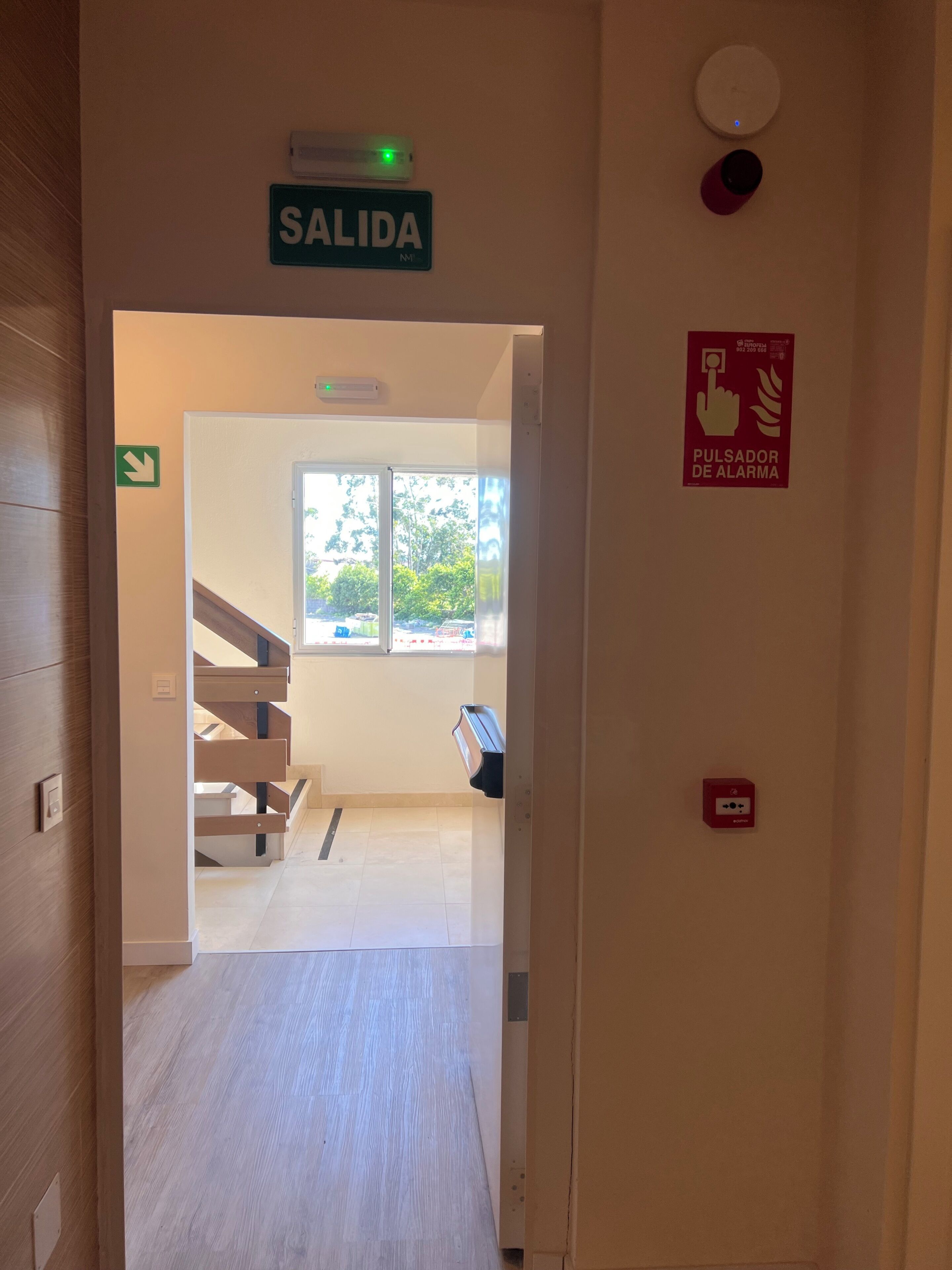 Un pasillo con un letrero que dice 'SALIDA', una ventana con vista a follaje, y un dispositivo de alarma contra incendios en la pared.