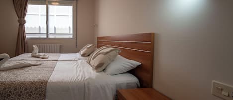 A hotel room with a bed, a wooden nightstand, a window with curtains, and a radiator.