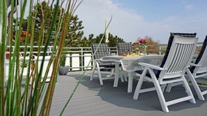En terrasse med hvide stole og et bord, omgivet af et hvidt hegn og grøn vegetation.