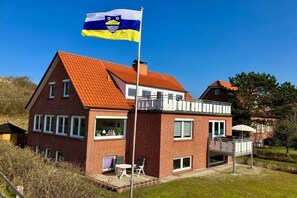 A two-story brick house with a red roof, a balcony, and a flagpole with a flag. - Amber in the House Sea Greeting (Juist)