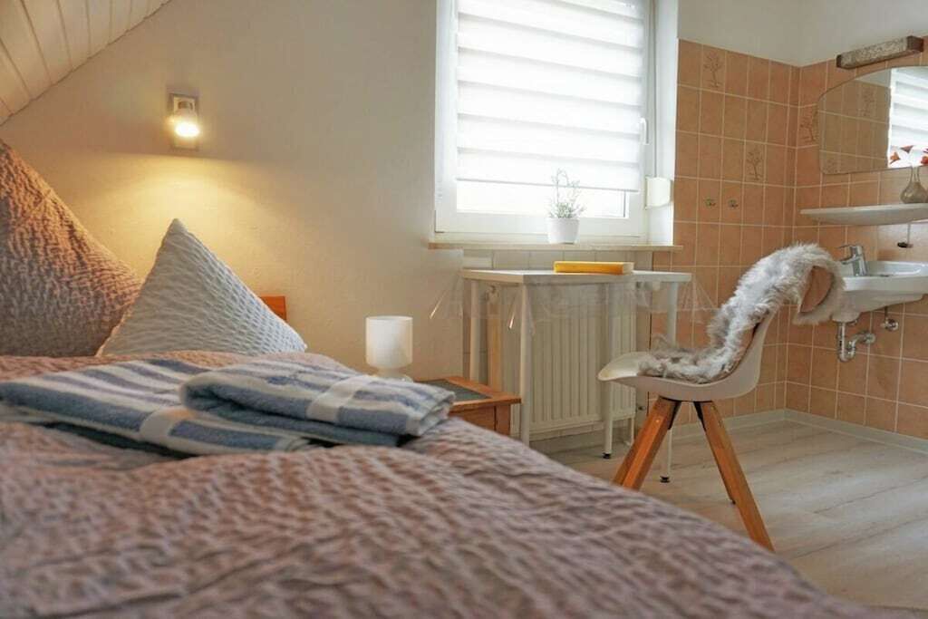 Quarto com cama, almofadas, mesa de cabeceira com abajur, janela com persianas, radiador e banheiro com pia e cadeira.