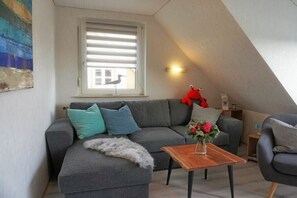 A cozy living room with a grey sofa, teal and grey cushions, a wooden coffee table, and a window with blinds. - Amber in the House Sea Greeting (Juist)