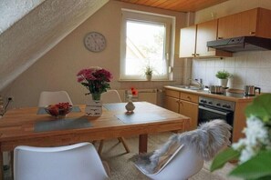 A cozy kitchen with wooden cabinets, a dining table with chairs, a clock on the wall, and a window providing natural light. - Amber in the House Sea Greeting (Juist)
