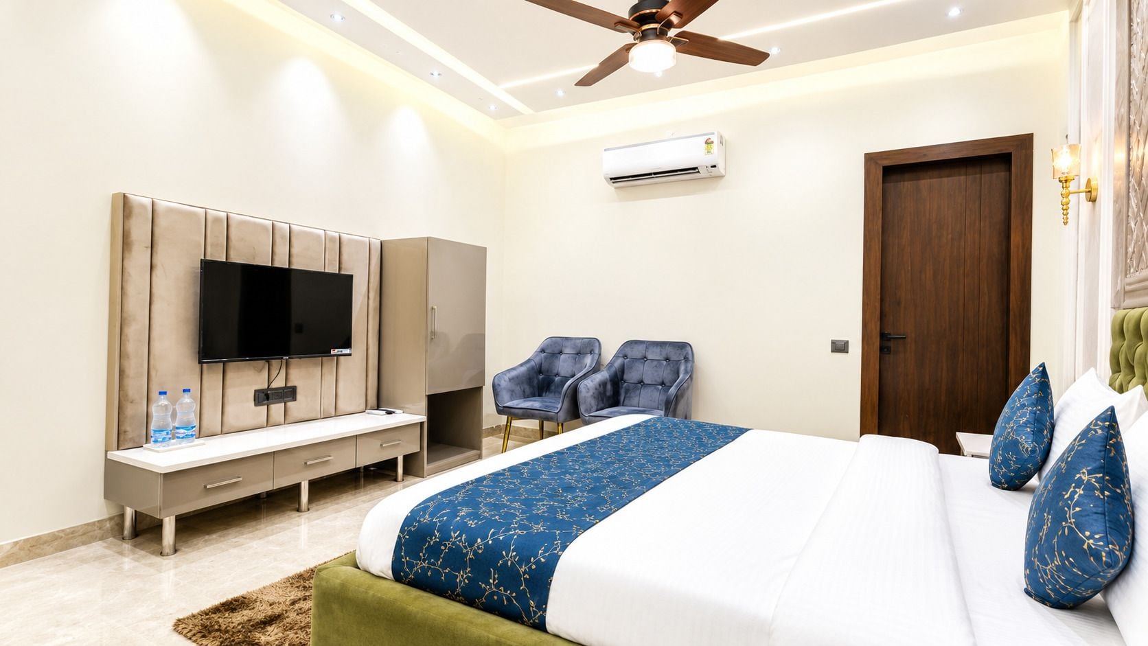 A hotel room with a bed, a TV, a wardrobe, two chairs, and an air conditioning unit.