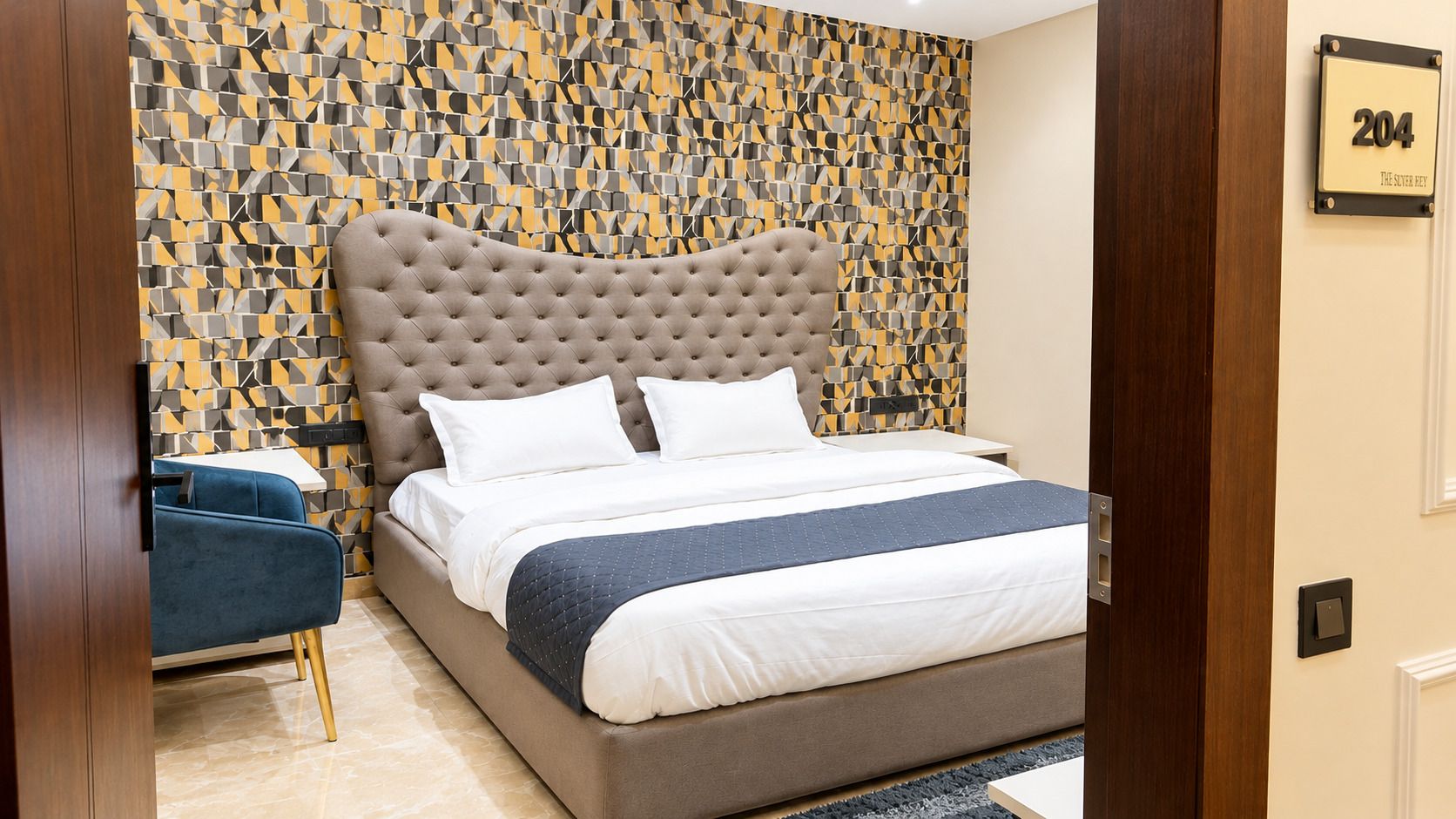 A hotel room with a tufted headboard, a bed with white linens, a blue chair, and a patterned wall.