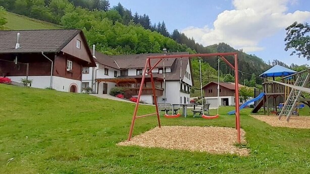 Un parque infantil con columpios y toboganes, rodeado de casas y una colina.