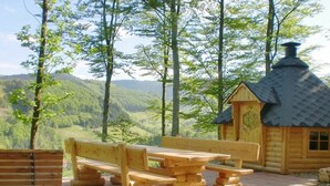 Una cabaña de madera con chimenea, rodeada por un banco y una mesa de picnic, con vista a un valle boscoso.