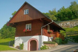 Ein zweistöckiges Holzhaus mit geneigtem Dach, einer Veranda mit Geländer und einer Steinsäule im Hintergrund.