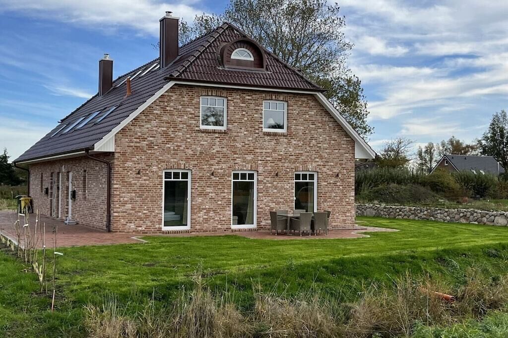Una casa de ladrillo de dos pisos con techo inclinado, un patio con mobiliario exterior y un césped bien cuidado.
