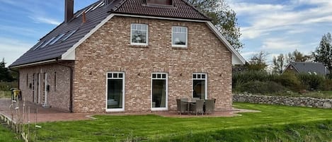 Ein zweigeschossiges Backsteinhhaus mit einem geneigten Dach, einer Terrasse mit Gartenmöbeln und einem gepflegten Rasen.